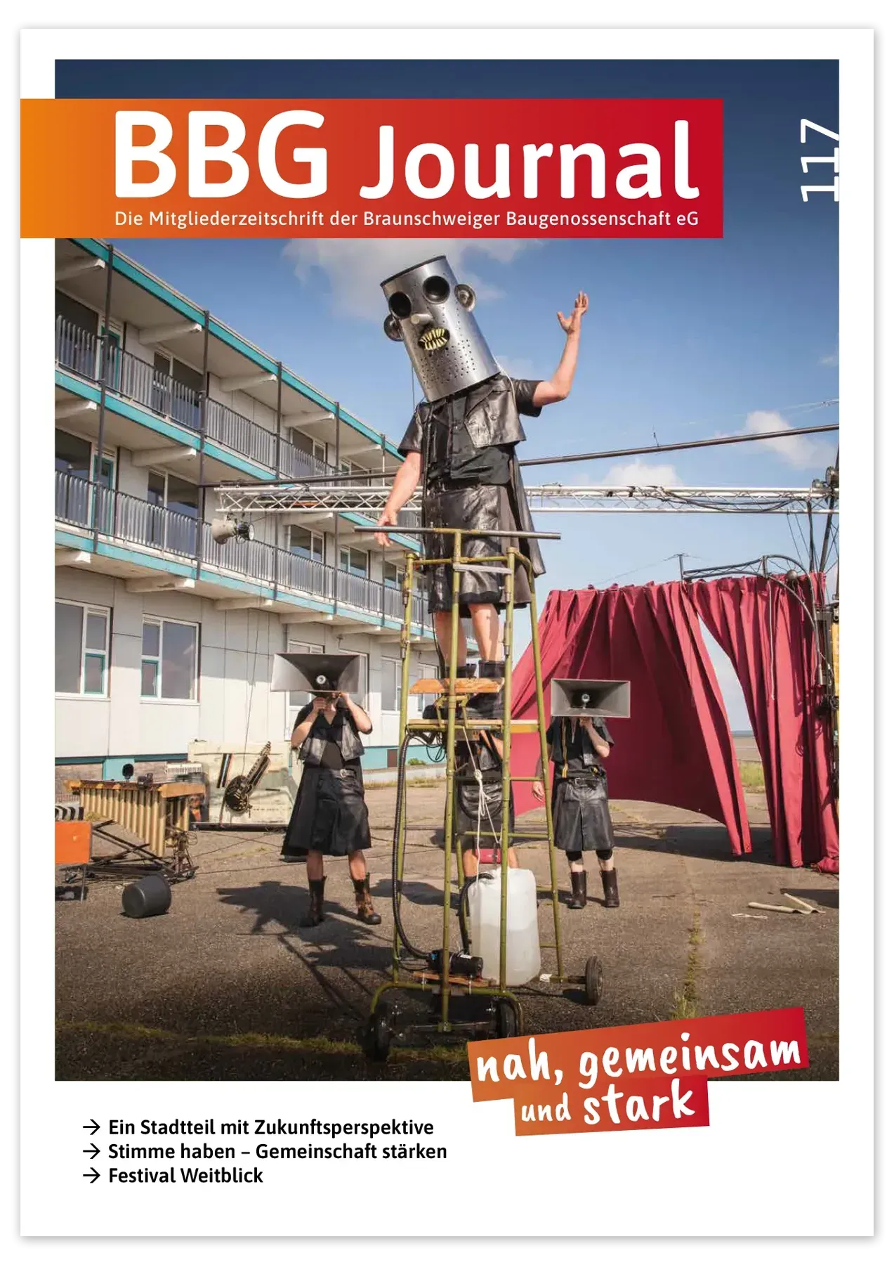 Drei Personen in schwarzen Kostümen und mit Metallhelmen treten im Freien auf einer hohen Metallkonstruktion mit roten Vorhängen und Wohnblocks im Hintergrund auf.