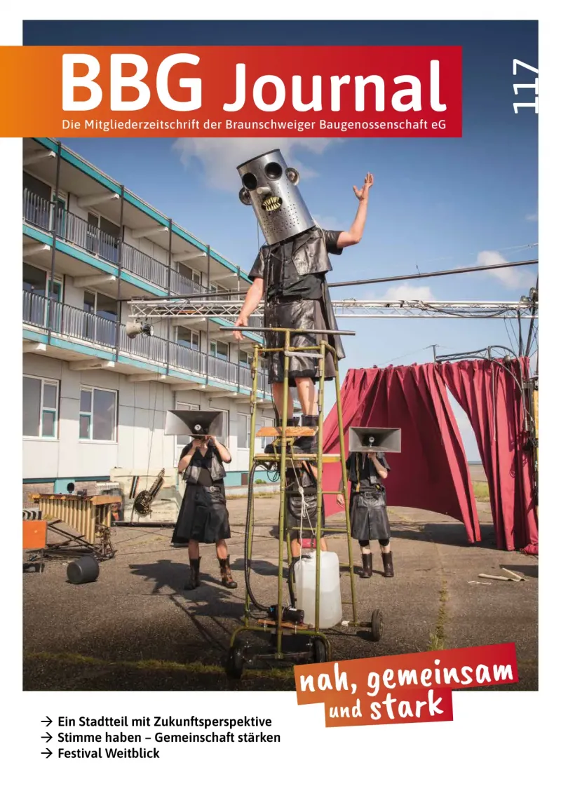 Drei Personen in schwarzer Kleidung und mit großen Metallroboterhelmen treten im Freien in der Nähe der roten Gebäude auf, einer von ihnen hebt die Arme.