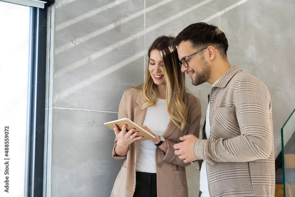 Zwei junge Erwachsene lächeln und schauen gemeinsam auf ein Tablet in einem modernen Innenraum.