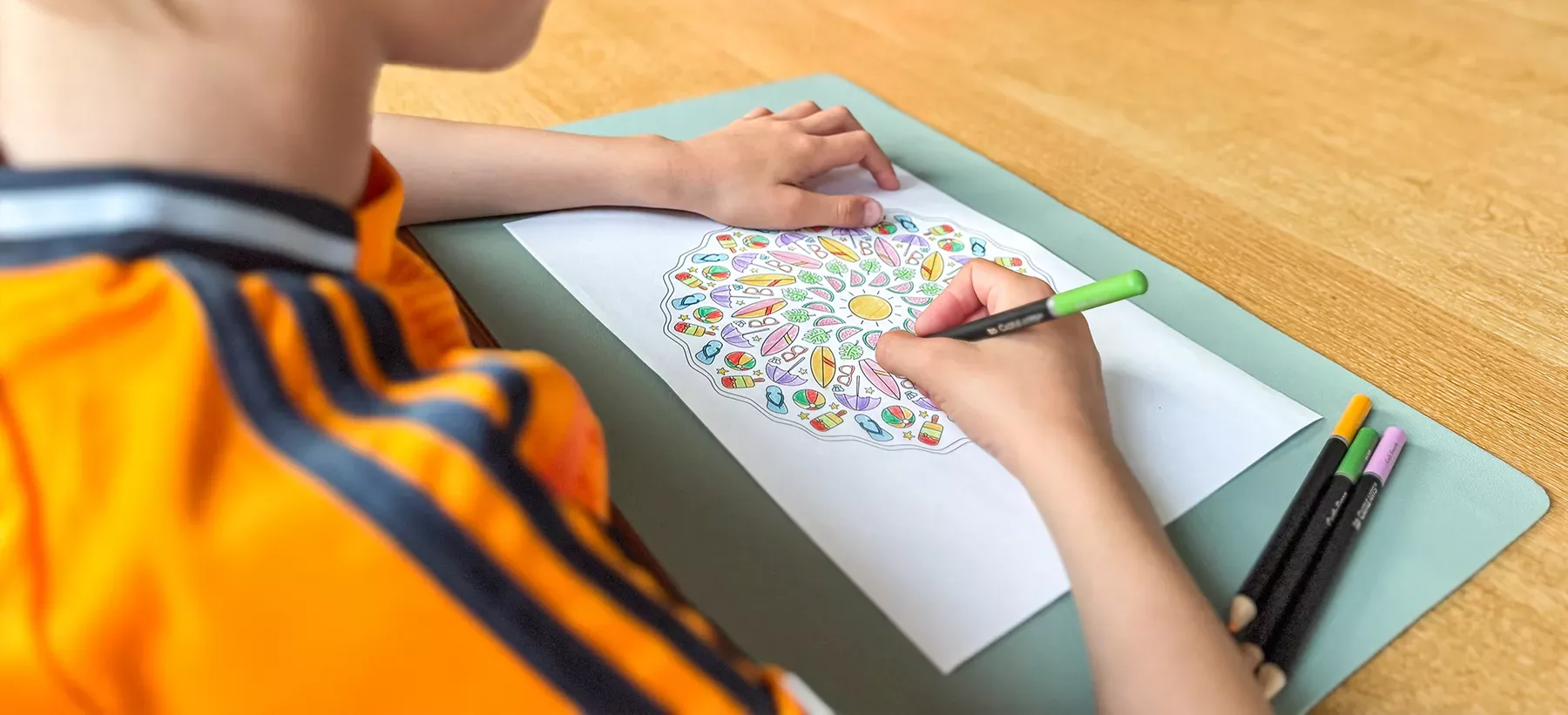 Ein Kind in einem orangefarbenen T-Shirt malt ein detailliertes Mandala mit Buntstiften auf ein weißes Blatt Papier auf einem Tisch.
