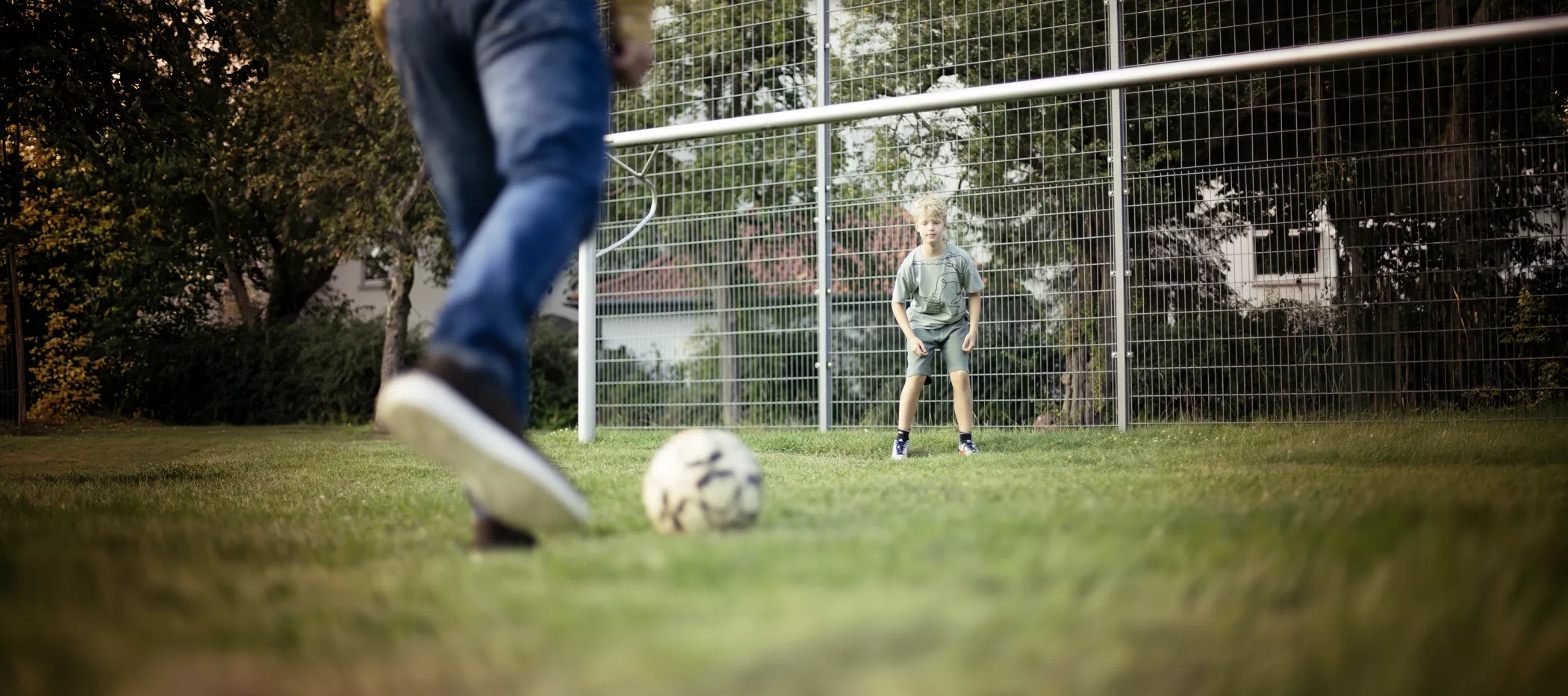 Ein Erwachsener macht sich bereit, einen Fußball in Richtung eines Kindes zu schießen, das auf einem Rasenplatz in der Torwartposition vor dem Tor steht.