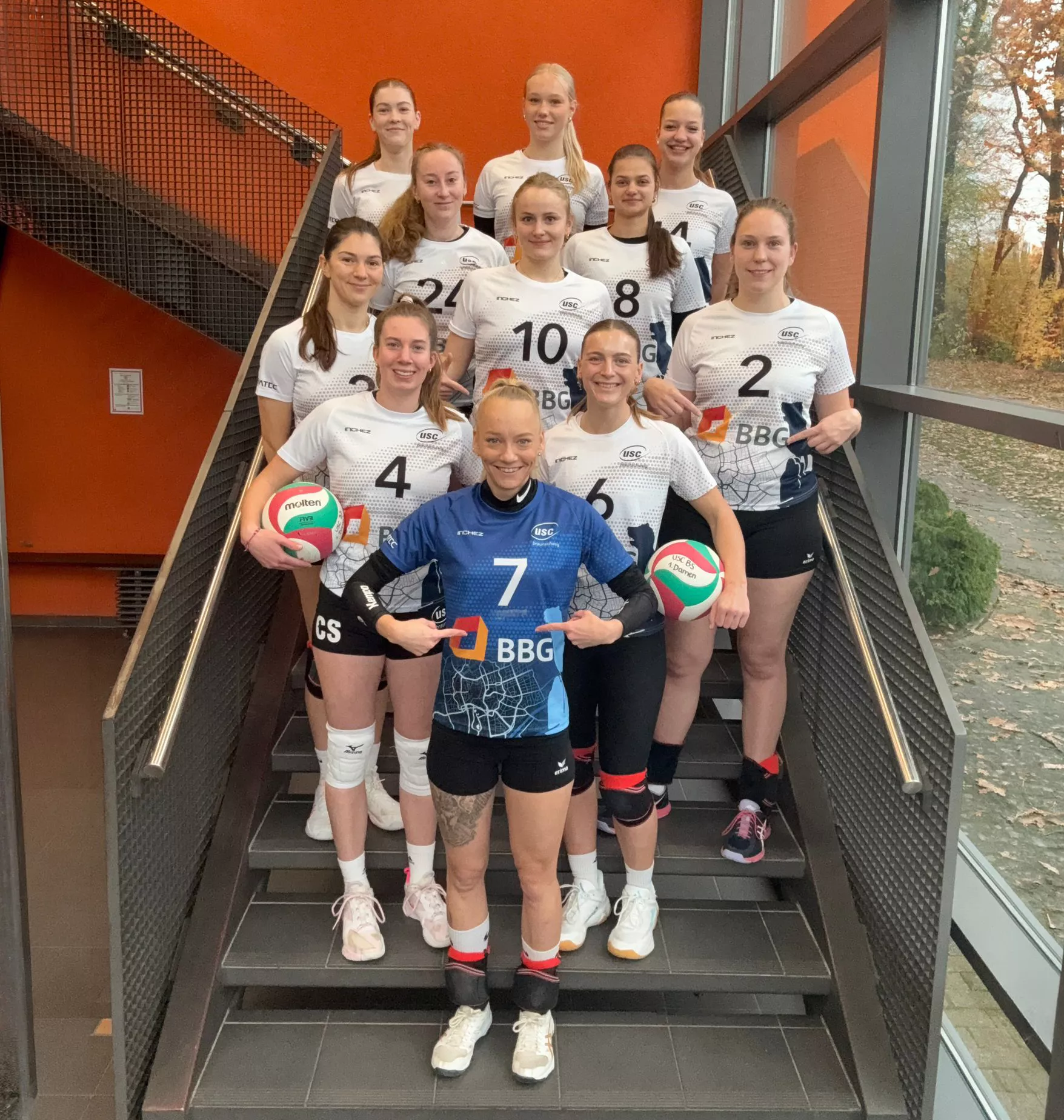 Zehn Frauen in Volleyball-Trikots stehen auf einer Treppe in einem Gebäude und halten Volleyballbälle in den Händen; im Hintergrund sind orangefarbene Wände mit Fenstern zu sehen.