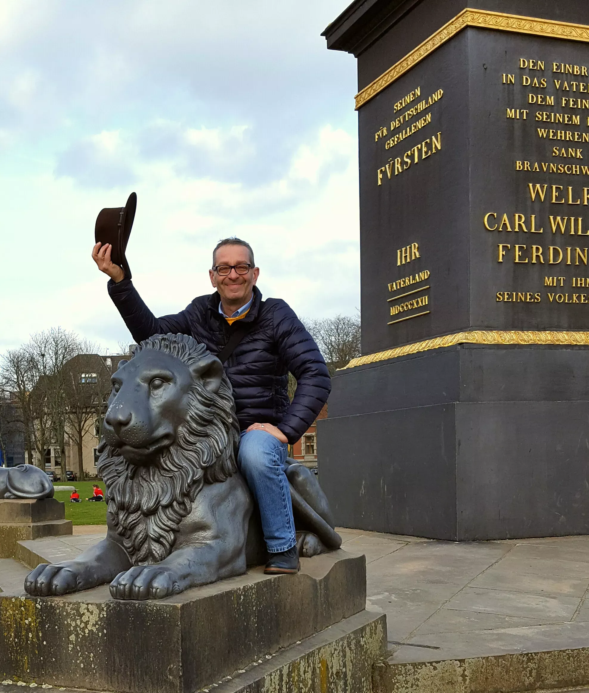 Ein lächelnder Mann, der auf einer Löwenstatue sitzt und einen Hut in die Luft hält, neben einem Denkmal mit goldener Schrift.