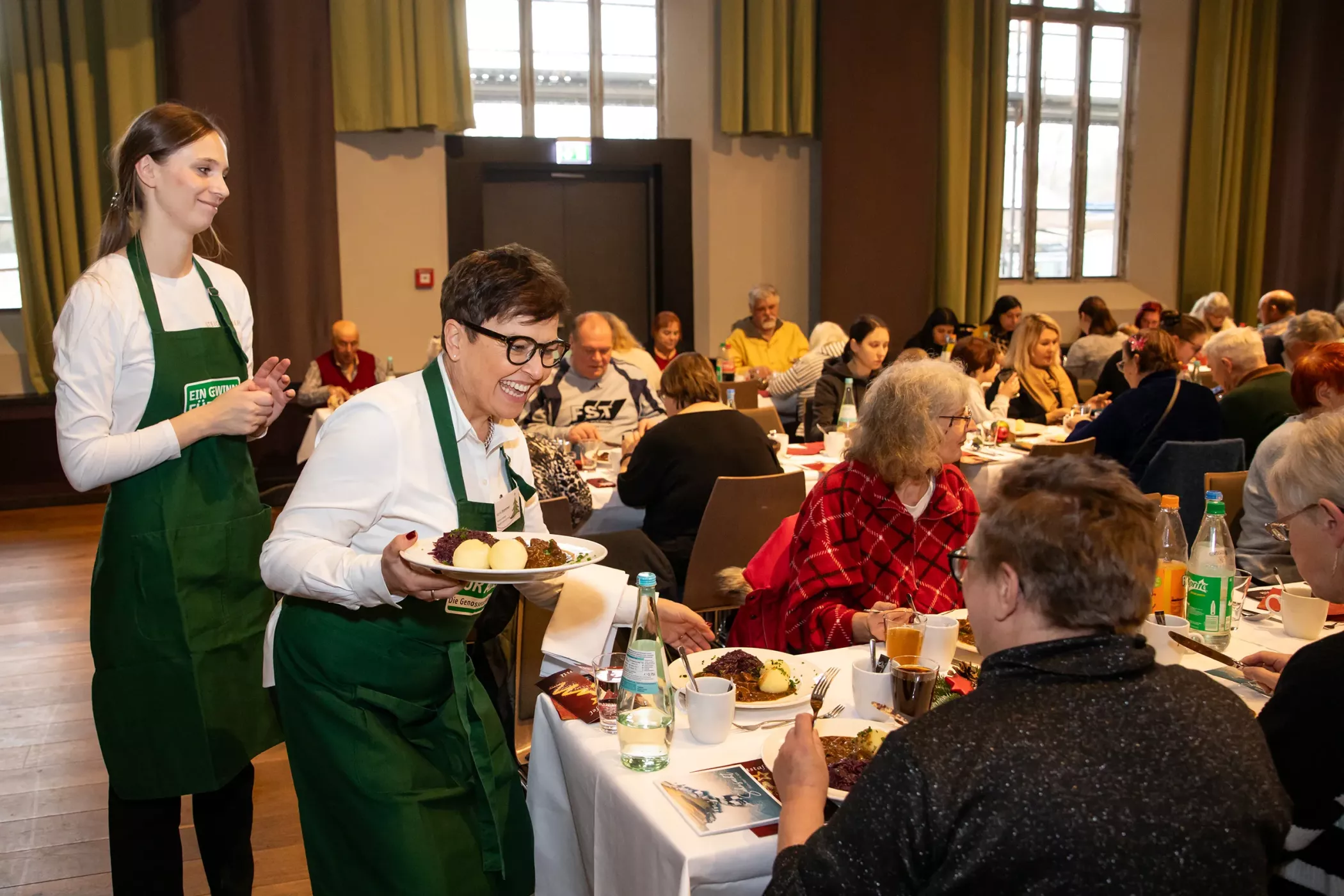 A woman in a green apron serves food to the guests at the tables in a crowded, well-lit dining room.