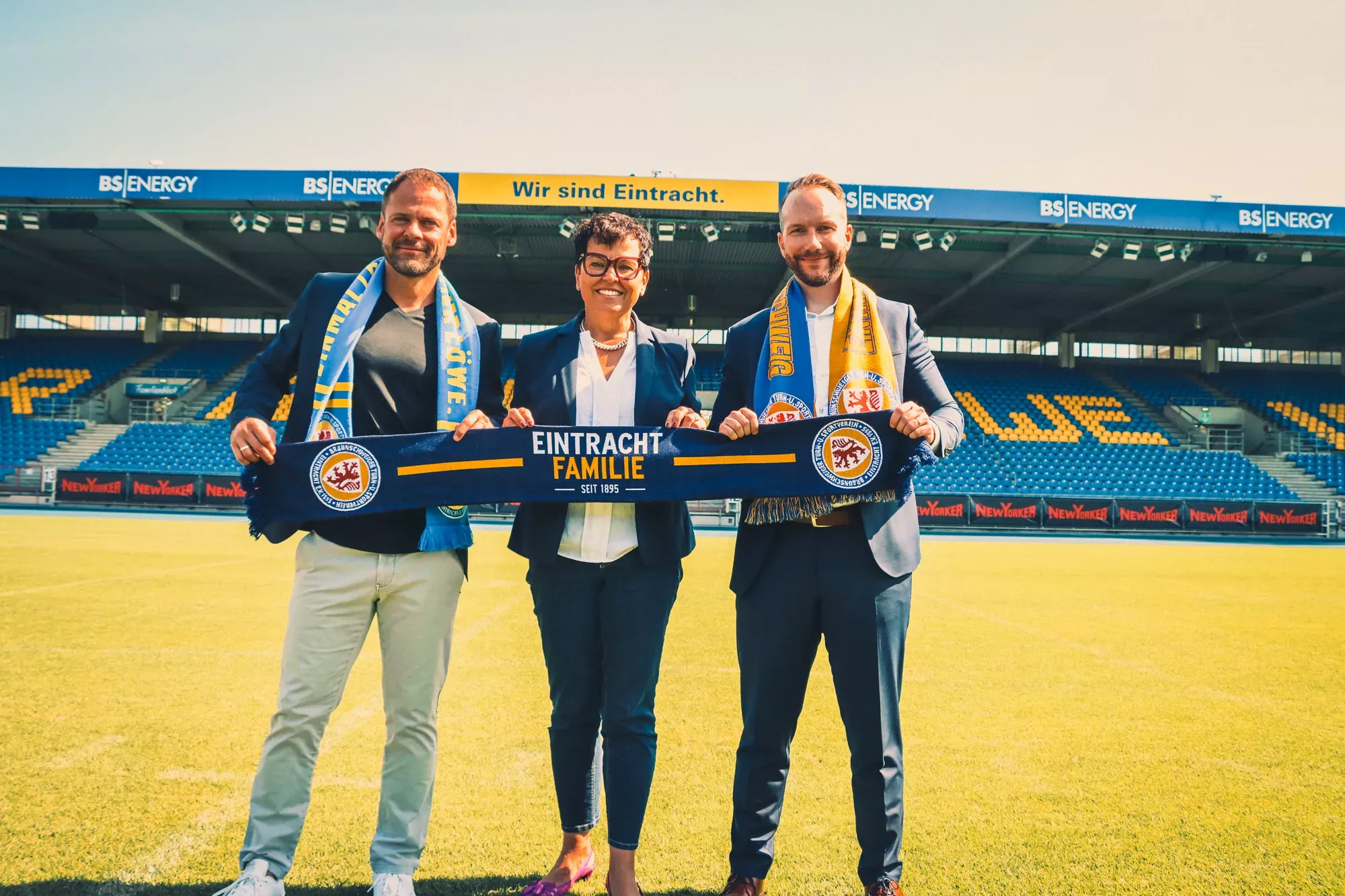 Three people standing in a soccer stadium, smiling and holding a Eintracht family scarf, with stadium seats and banners in the background.