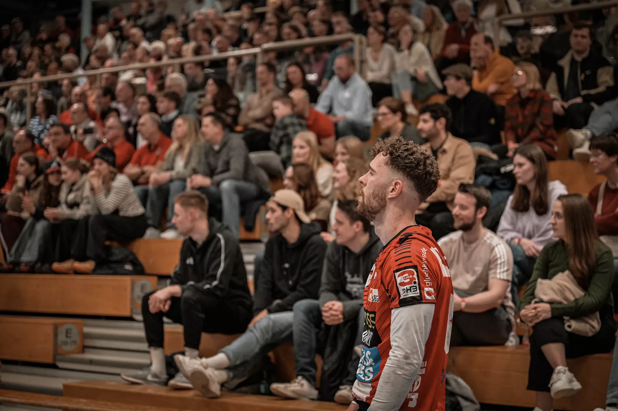 A man in a red sports shirt stands in front of a crowded grandstand with spectators sitting at an indoor event.