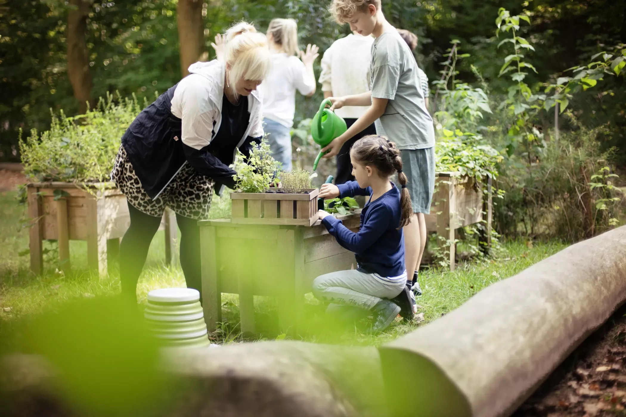 A group of people and children gardening together outdoors, surrounded by green plants and small wooden planters.