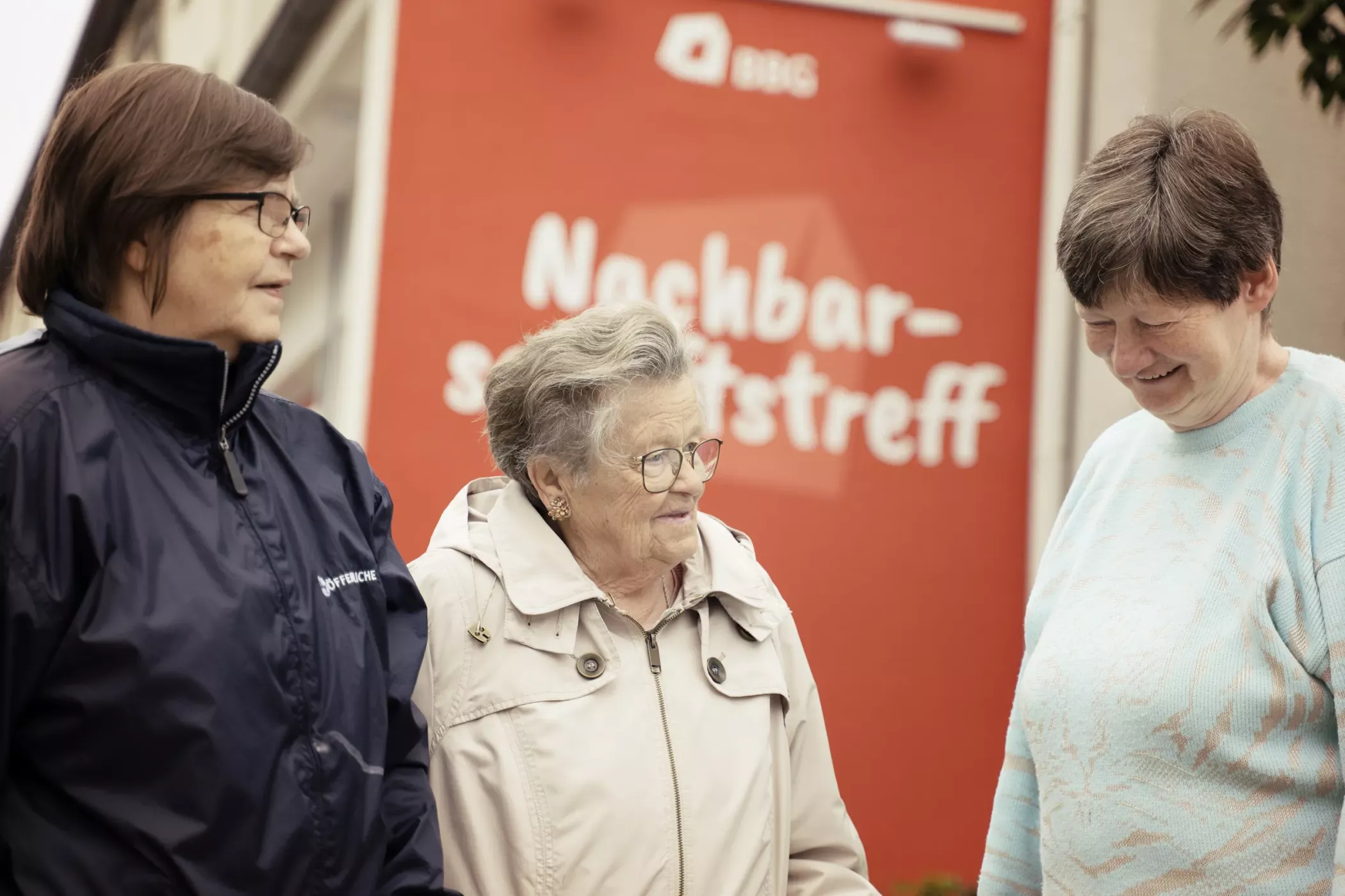 Üç yaşlı kadın, Nachbarschaftstreff'i (komşular için buluşma yeri) gösteren kırmızı bir tabelanın önünde gülümseyerek duruyor.