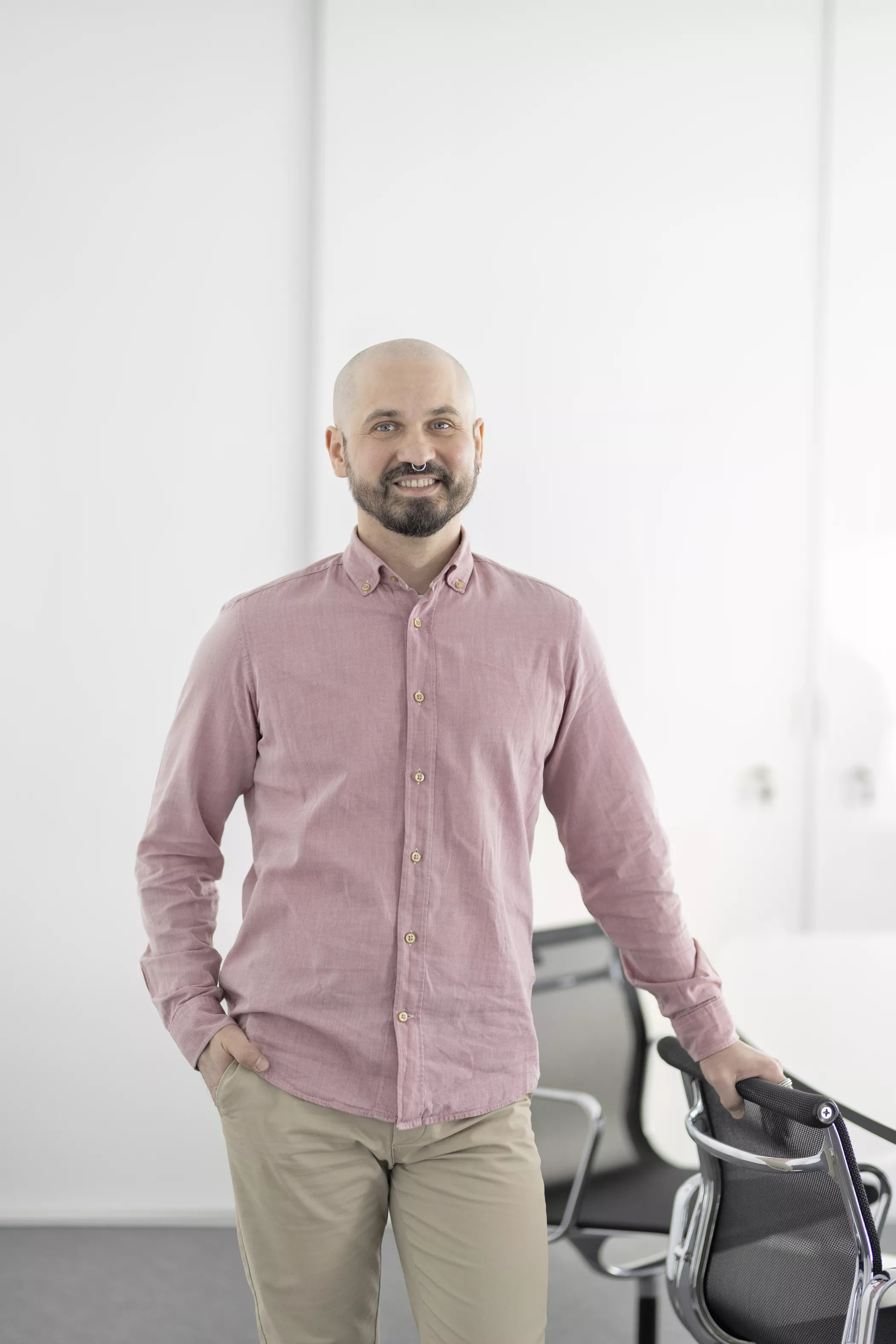 Ein lächelnder, bärtiger Mann in einem rosa Hemd steht neben einem Stuhl in einem hellen, modernen Besprechungsraum.