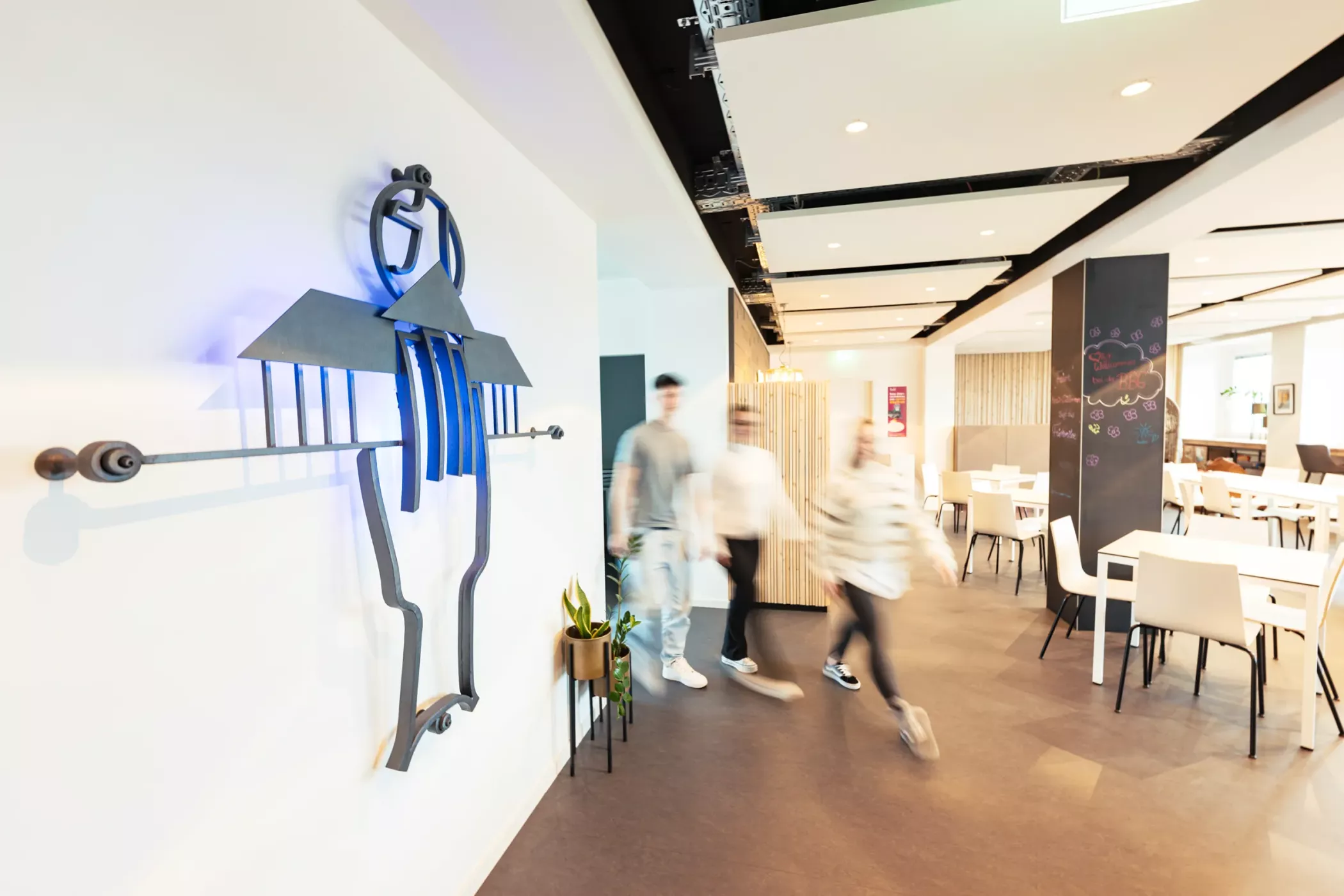 Trois personnes entrent dans une cafétéria moderne et lumineuse, avec des tables blanches et des peintures murales abstraites sur la gauche.