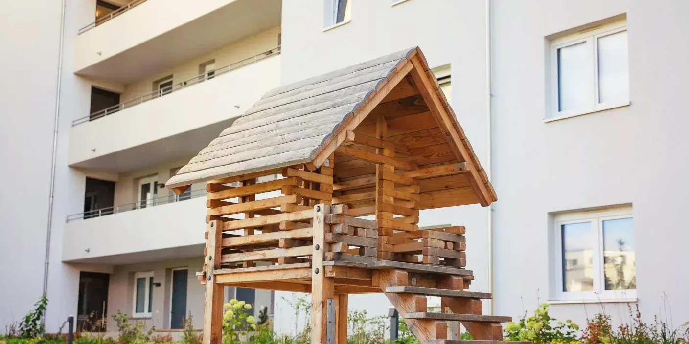 Ein kleines Holzspielhaus auf Stelzen mit Treppe vor einem modernen Wohnblock.