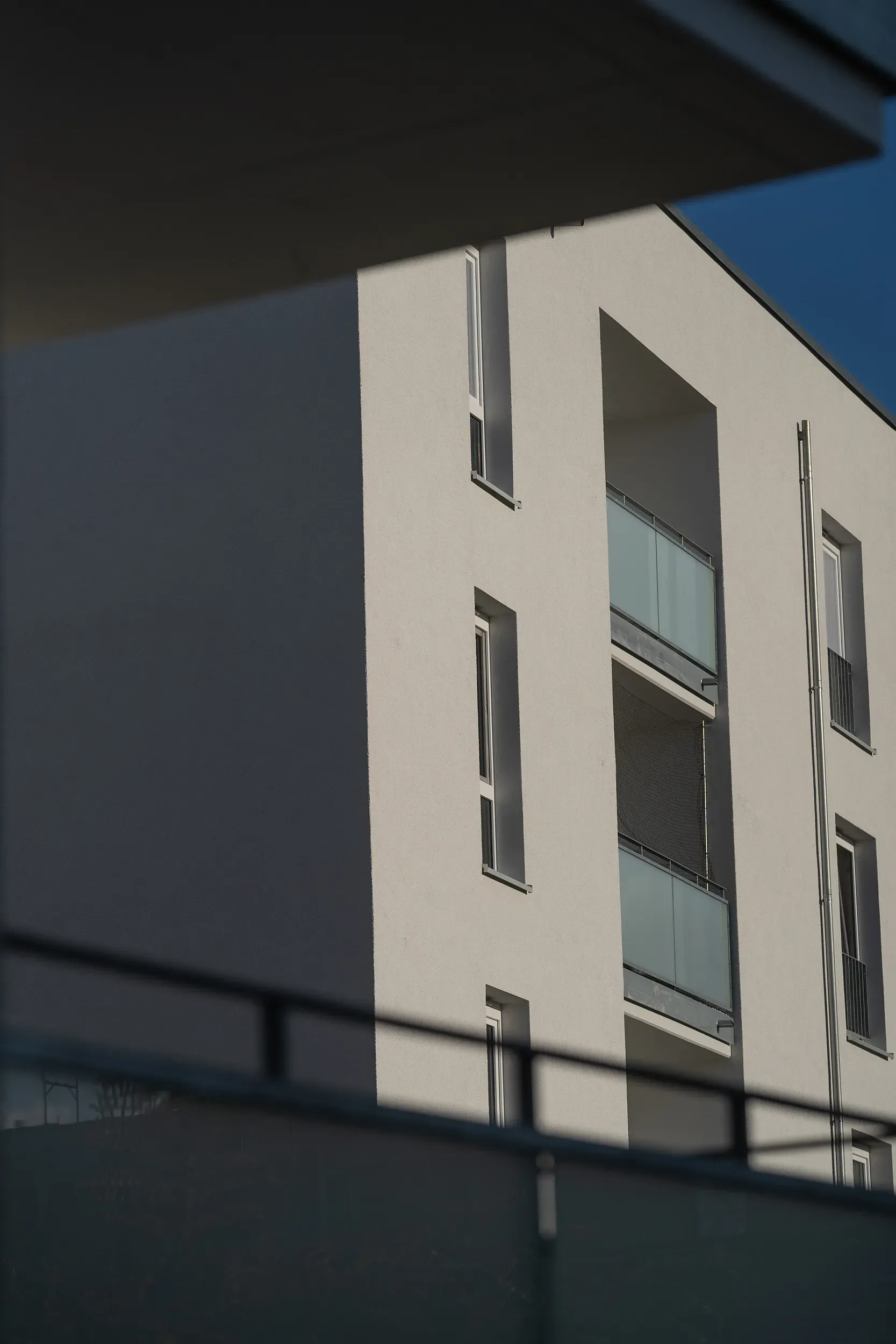 A modern white residential building with partially shaded glass balconies against a clear blue sky.