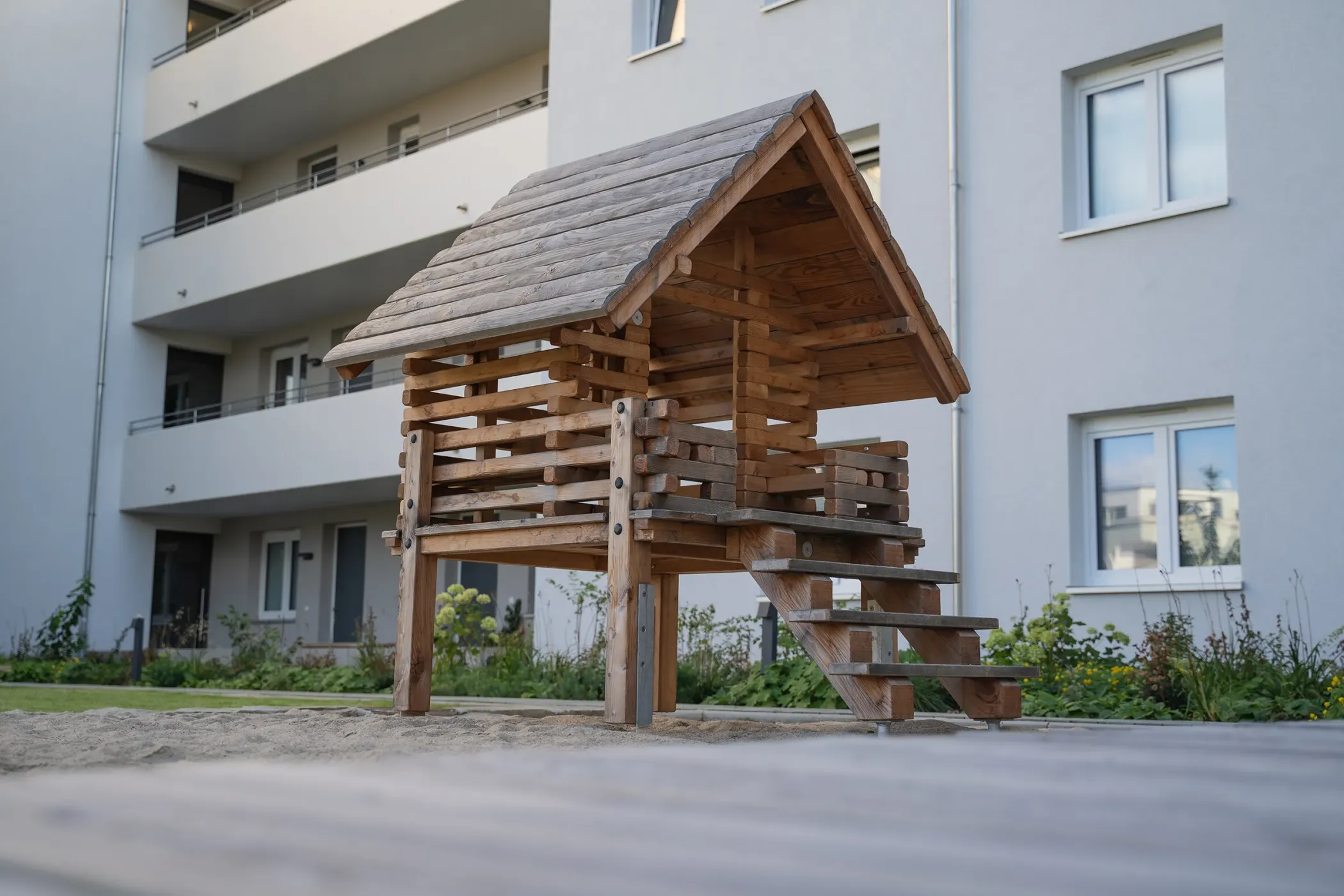A small wooden playhouse with stairs stands in front of a modern block of flats.