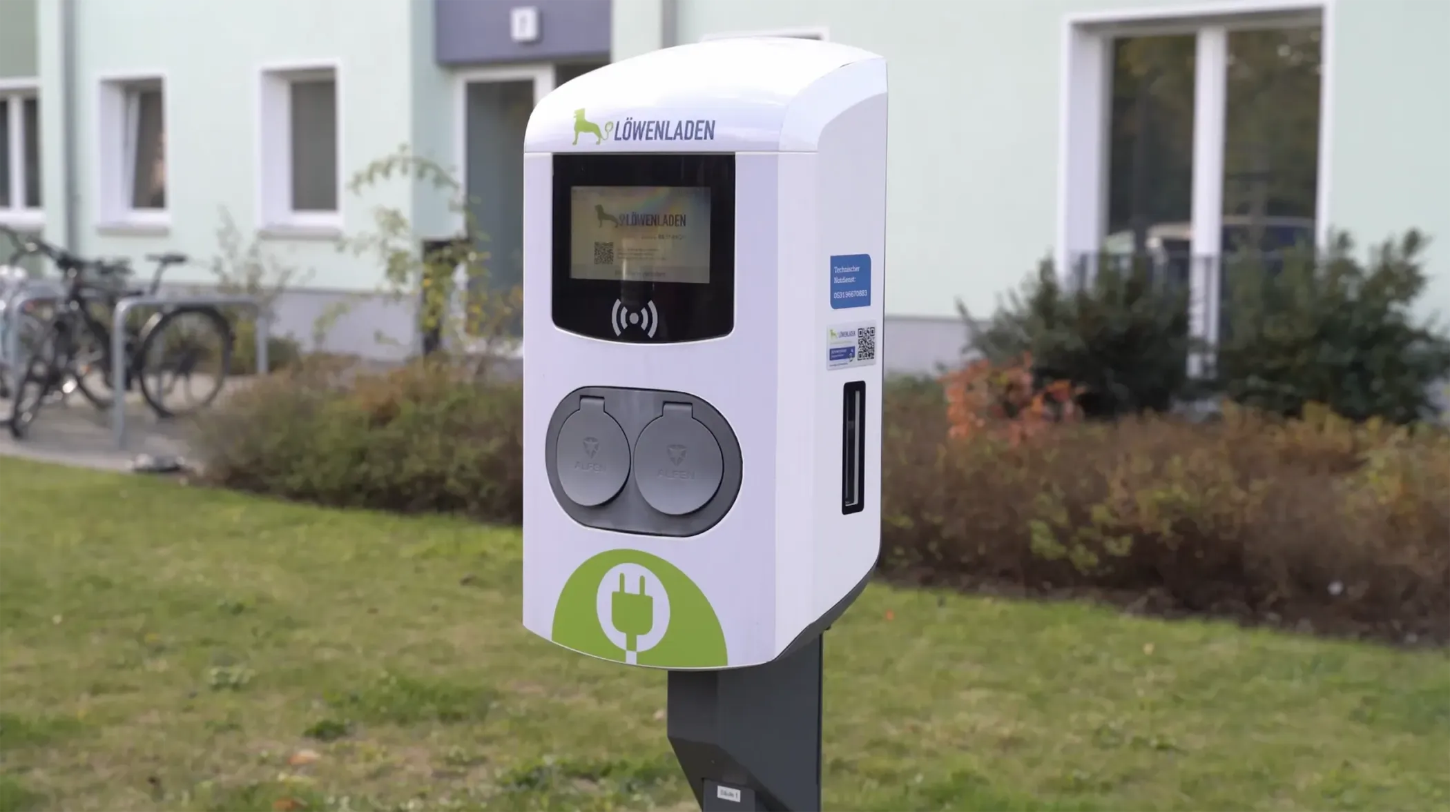 A charging station for electric vehicles with two charging connections, installed outdoors on the lawn in front of a building.