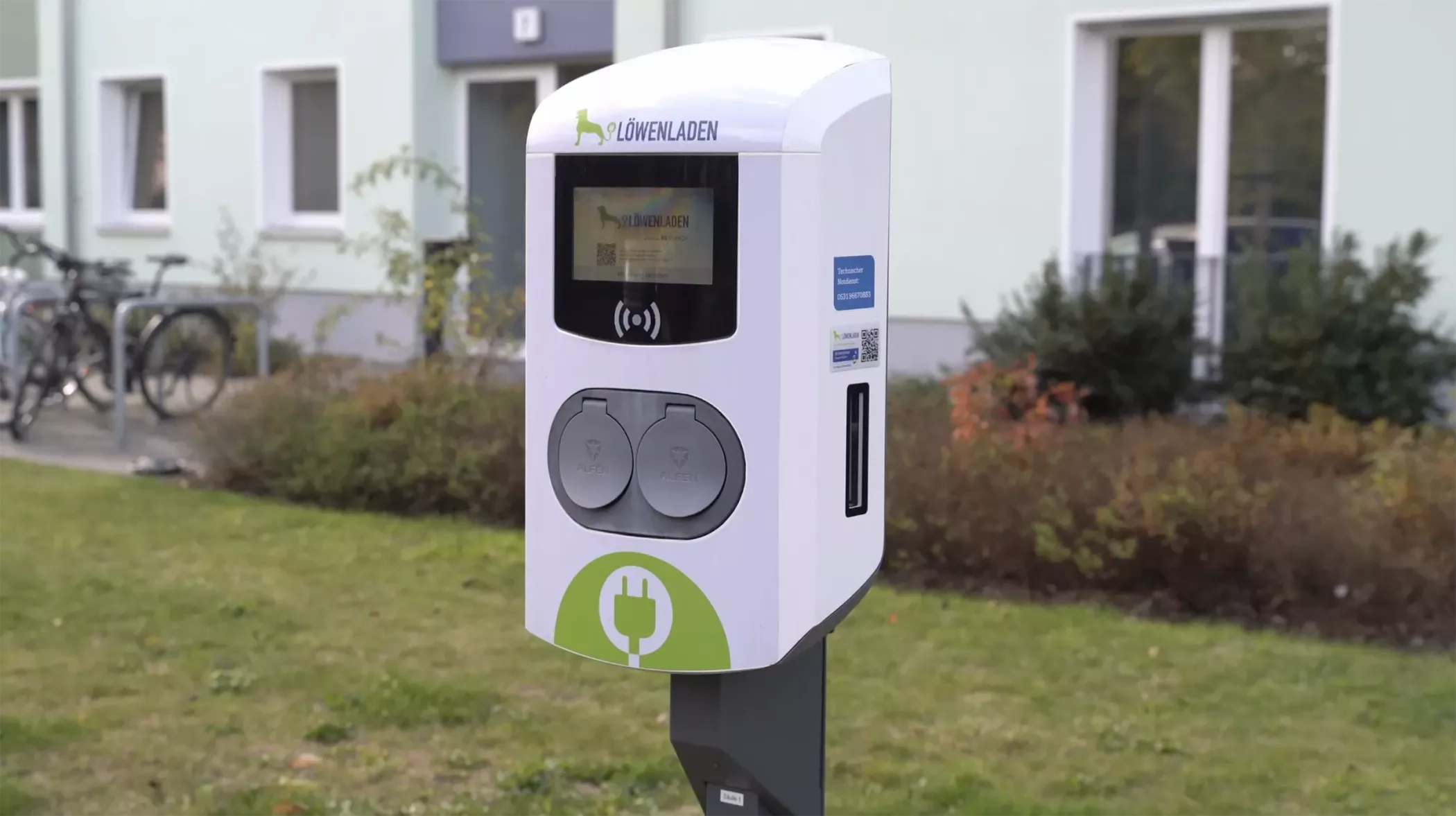 Eine Ladestation für Elektrofahrzeuge mit zwei Ladeanschlüssen, die im Freien auf der Wiese vor einem Gebäude installiert ist.