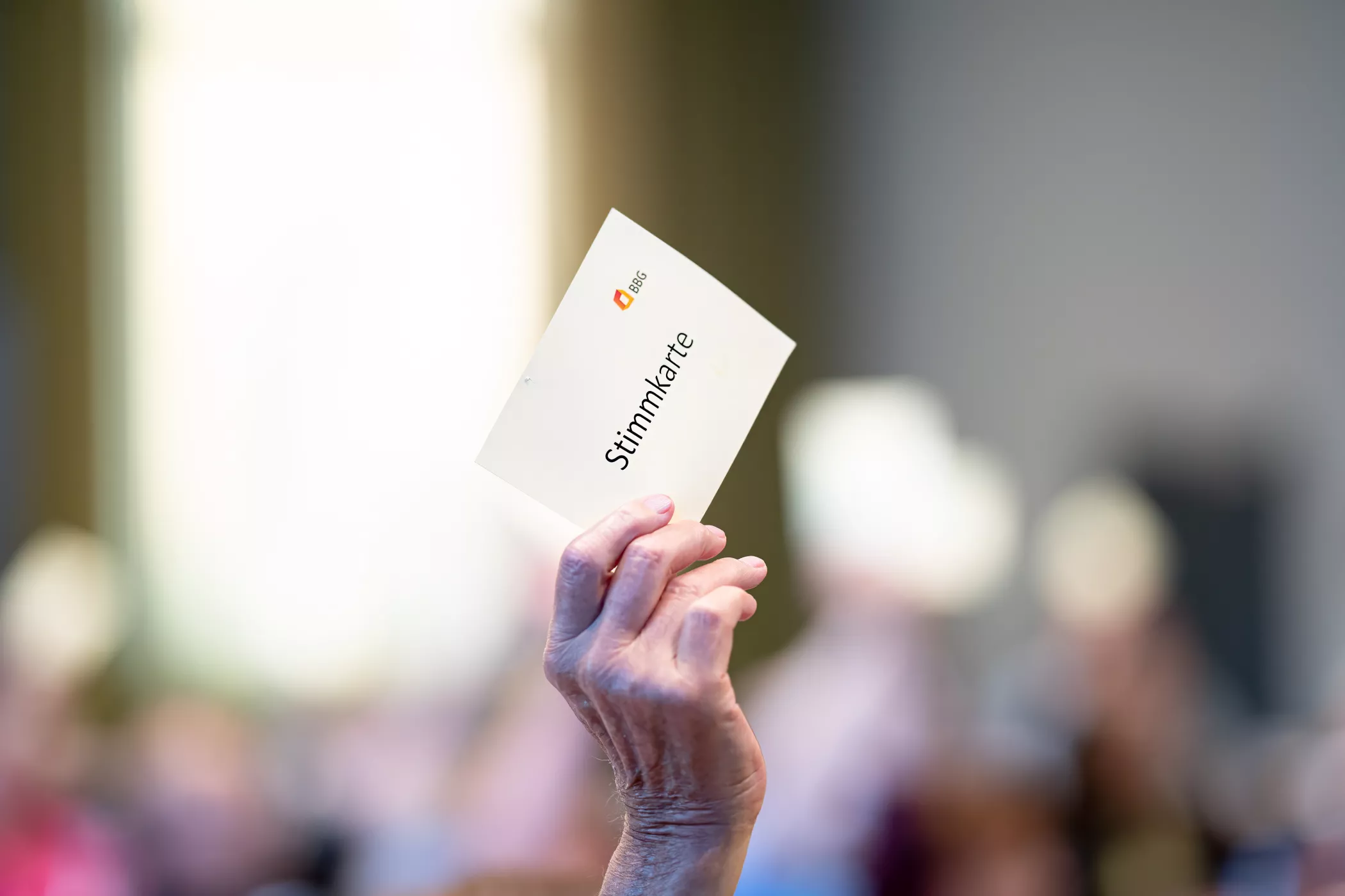 Eine Hand hält eine Wahlkarte mit der Aufschrift Stimmkarte in einem überfüllten, verschwommenen Raum.