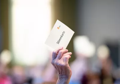 Eine Hand hält eine Wahlkarte mit der Aufschrift Stimmkarte in einem überfüllten, verschwommenen Raum.