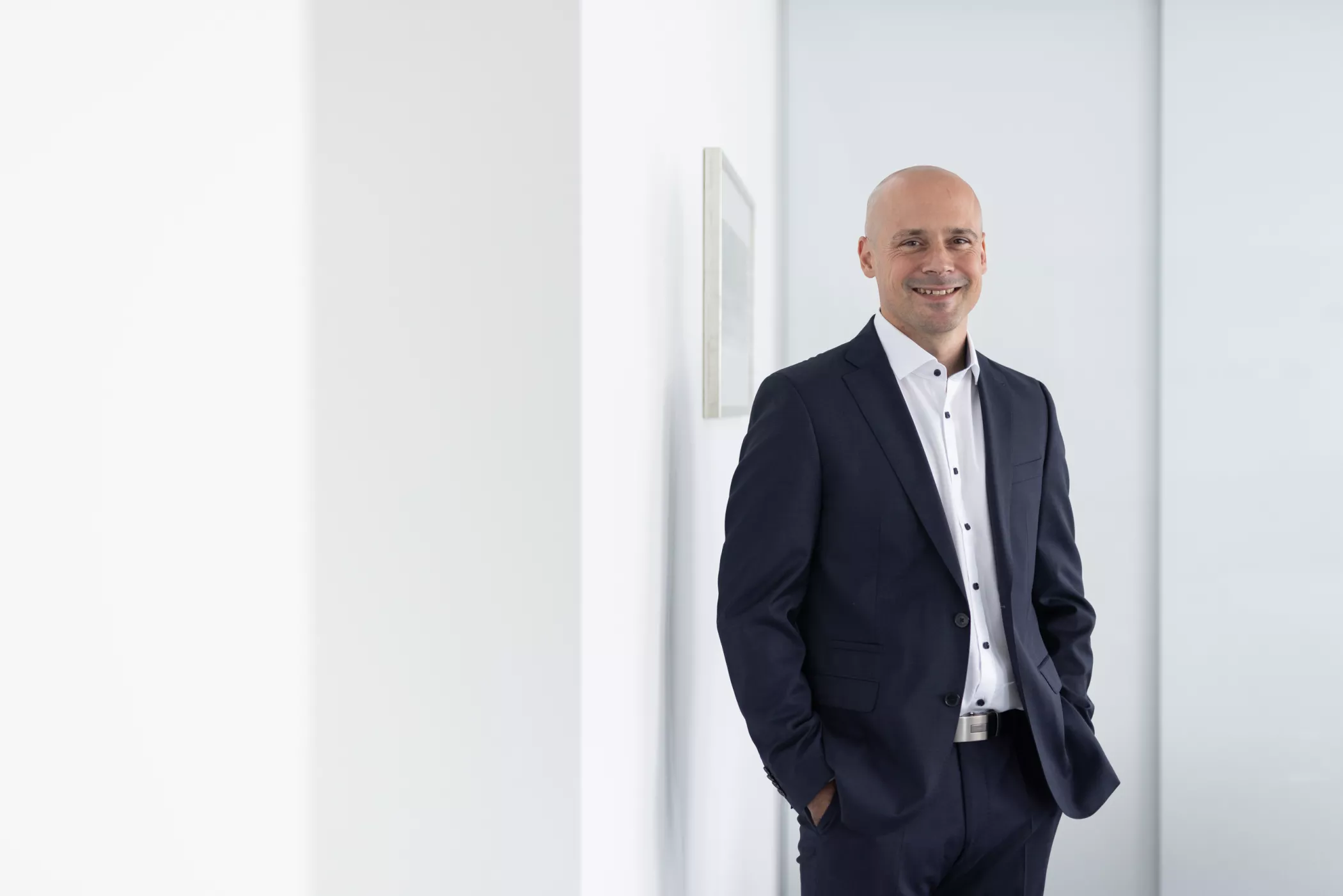 Ein glatzköpfiger Mann in einem dunklen Anzug und weißem Hemd steht lächelnd mit den Händen in den Taschen neben einer weißen Wand in einer hellen, modernen Umgebung.