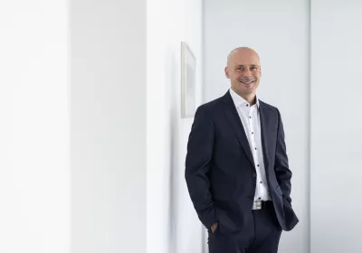 Ein glatzköpfiger Mann in einem dunklen Anzug und weißem Hemd steht lächelnd mit den Händen in den Taschen neben einer weißen Wand in einer hellen, modernen Umgebung.