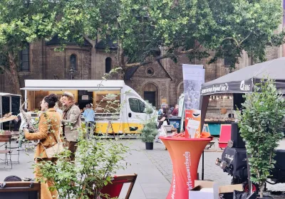 Eine Marktszene im Freien mit Menschen, die sich unterhalten, Ständen, einem Imbisswagen und Grünflächen auf dem Stadtplatz in der Nähe eines alten Steingebäudes.