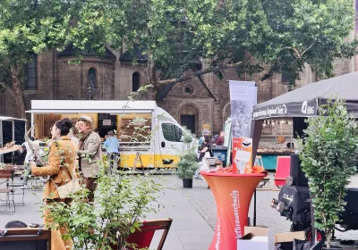 Eine Marktszene im Freien mit Menschen, die sich unterhalten, Ständen, einem Imbisswagen und Grünflächen auf dem Stadtplatz in der Nähe eines alten Steingebäudes.