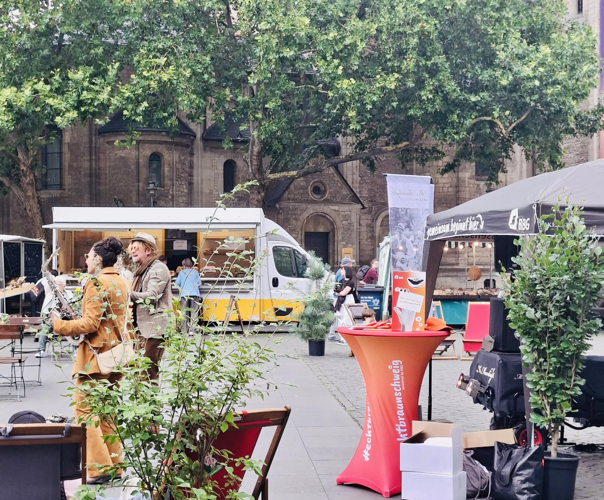 Eine Marktszene im Freien mit Menschen, die sich unterhalten, Ständen, einem Imbisswagen und Grünflächen auf dem Stadtplatz in der Nähe eines alten Steingebäudes.