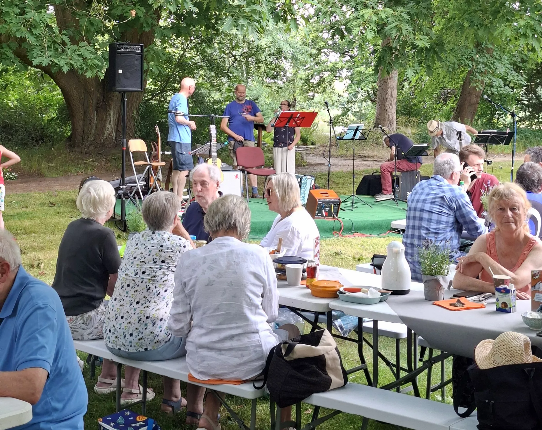 Die Menschen sitzen an Picknicktischen im Freien, plaudern und essen, während Musiker auf einer kleinen Bühne unter den Bäumen im Hintergrund ihre Instrumente aufstellen.