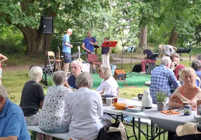 Die Menschen sitzen an Picknicktischen im Freien, plaudern und essen, während Musiker auf einer kleinen Bühne unter den Bäumen im Hintergrund ihre Instrumente aufstellen.