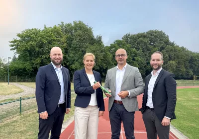 Vier Personen in Arbeitskleidung stehen auf der Laufbahn und lächeln, im Hintergrund sind Bäume und Gras zu sehen.