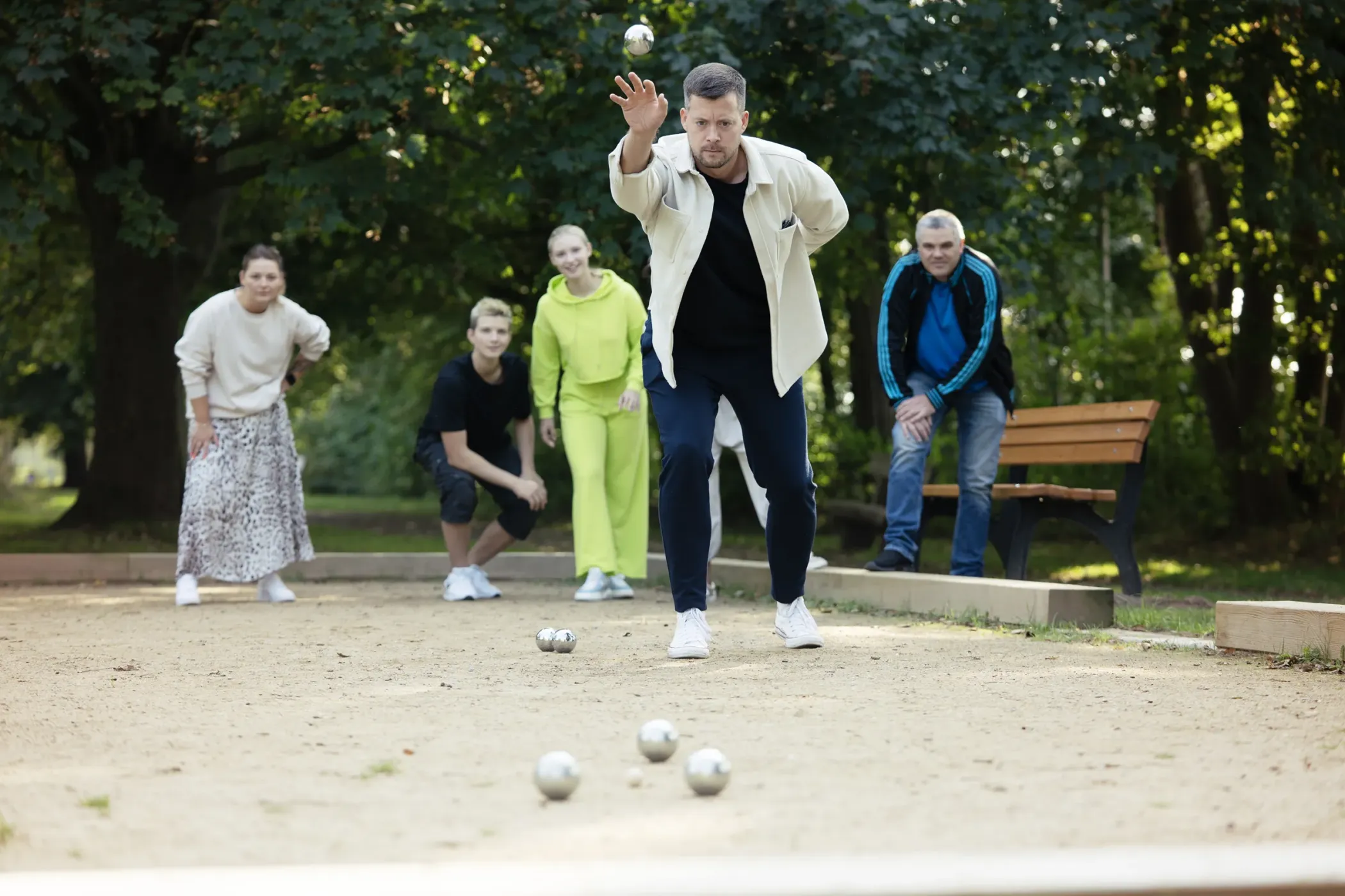 Ein Mann wirft eine Kugel bei einem Boulespiel, während fünf Personen in einem Park lächelnd zusehen.