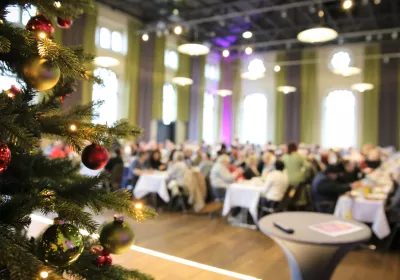 Im Mittelpunkt steht ein geschmückter Weihnachtsbaum, im Hintergrund eine große Gruppe von Menschen, die an Tischen in einem festlichen Saal sitzen.