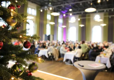 Im Mittelpunkt steht ein geschmückter Weihnachtsbaum, im Hintergrund eine große Gruppe von Menschen, die an Tischen in einem festlichen Saal sitzen.