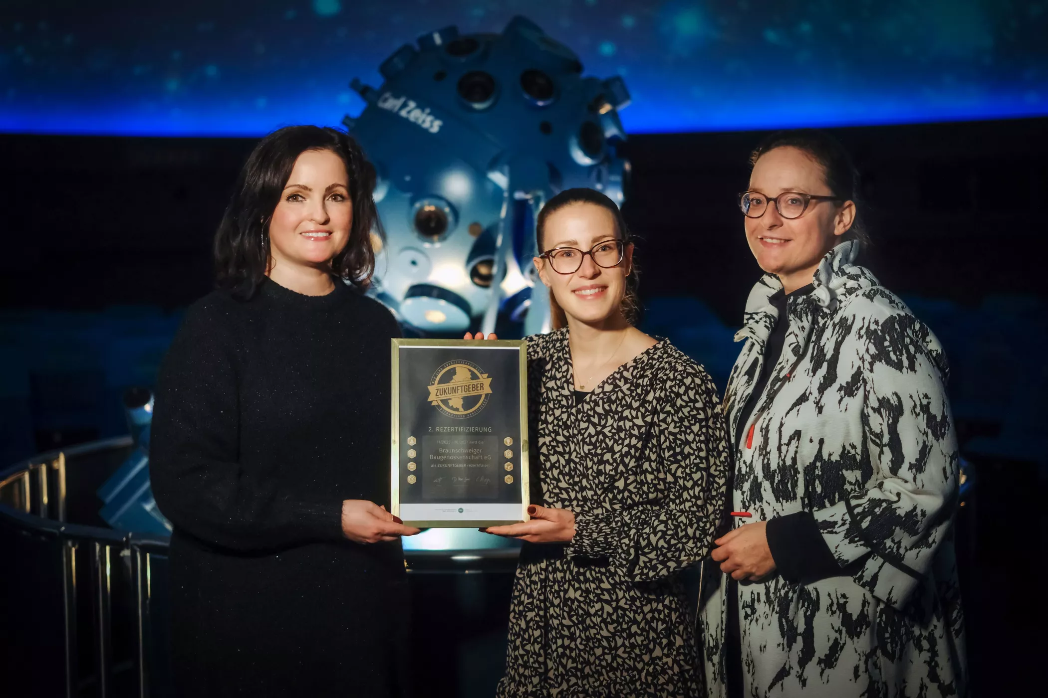 Drei Frauen stehen zusammen, lächeln und halten eine Plakette vor einem großen wissenschaftlichen Instrument.
