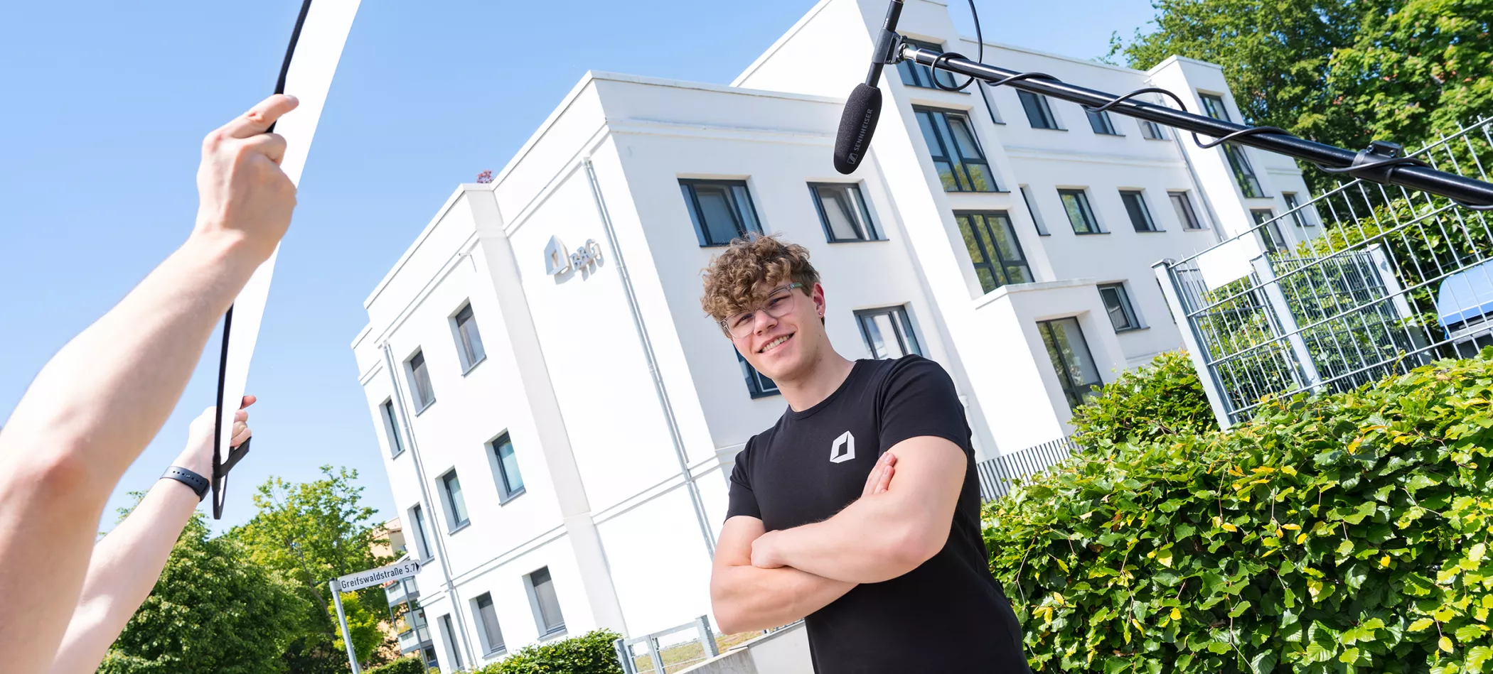Eine Person steht vor einem weißen Gebäude, die Arme verschränkt; neben ihr sind Aufnahmegeräte zu sehen, im Hintergrund sind grüne Pflanzen zu erkennen.
