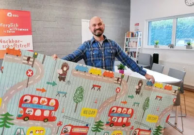 A smiling man holds a colorful play mat with cartoon vehicles and animals in a bright, modern room with bookshelves and posters.