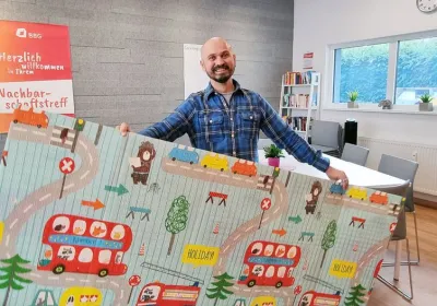 Ein lächelnder Mann hält eine bunte Spielmatte mit Cartoon-Fahrzeugen und -Tieren in einem hellen, modernen Raum mit Bücherregalen und Postern.