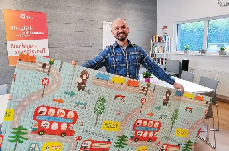 Ein lächelnder Mann hält eine bunte Spielmatte mit Cartoon-Fahrzeugen und -Tieren in einem hellen, modernen Raum mit Bücherregalen und Postern.