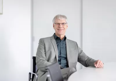 Ein Mann mit grauem Haar und Brille, der einen grauen Anzug trägt, sitzt lächelnd an einem weißen Tisch in einem hellen Büro.