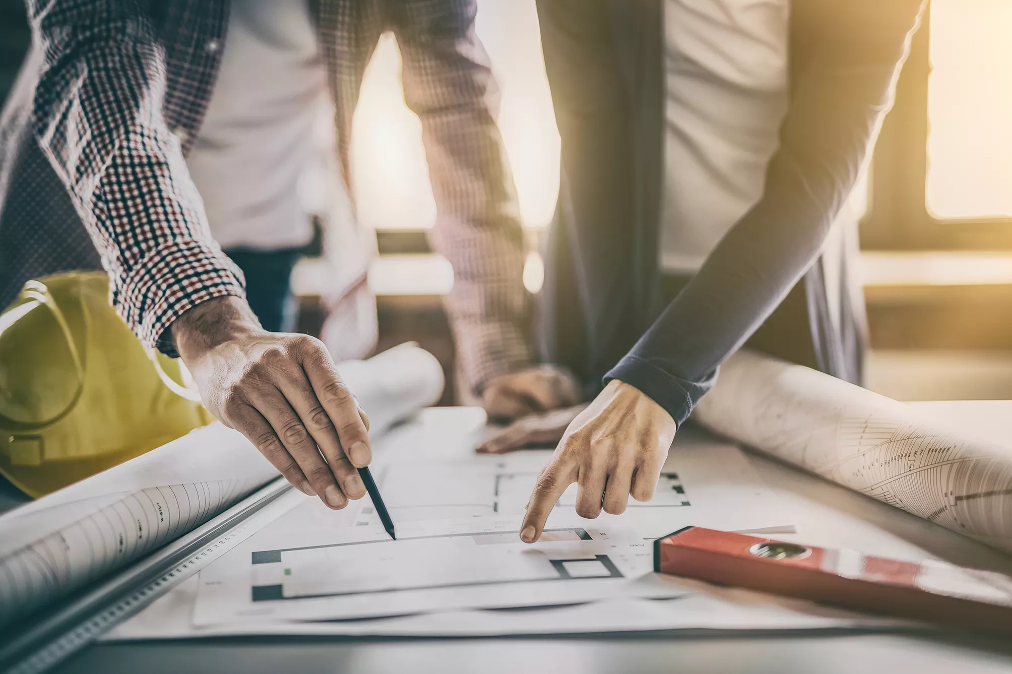 Zwei Personen, die auf Architekturpläne auf einem Tisch zeigen, umgeben von Zeichengeräten und einem gelben Hut.