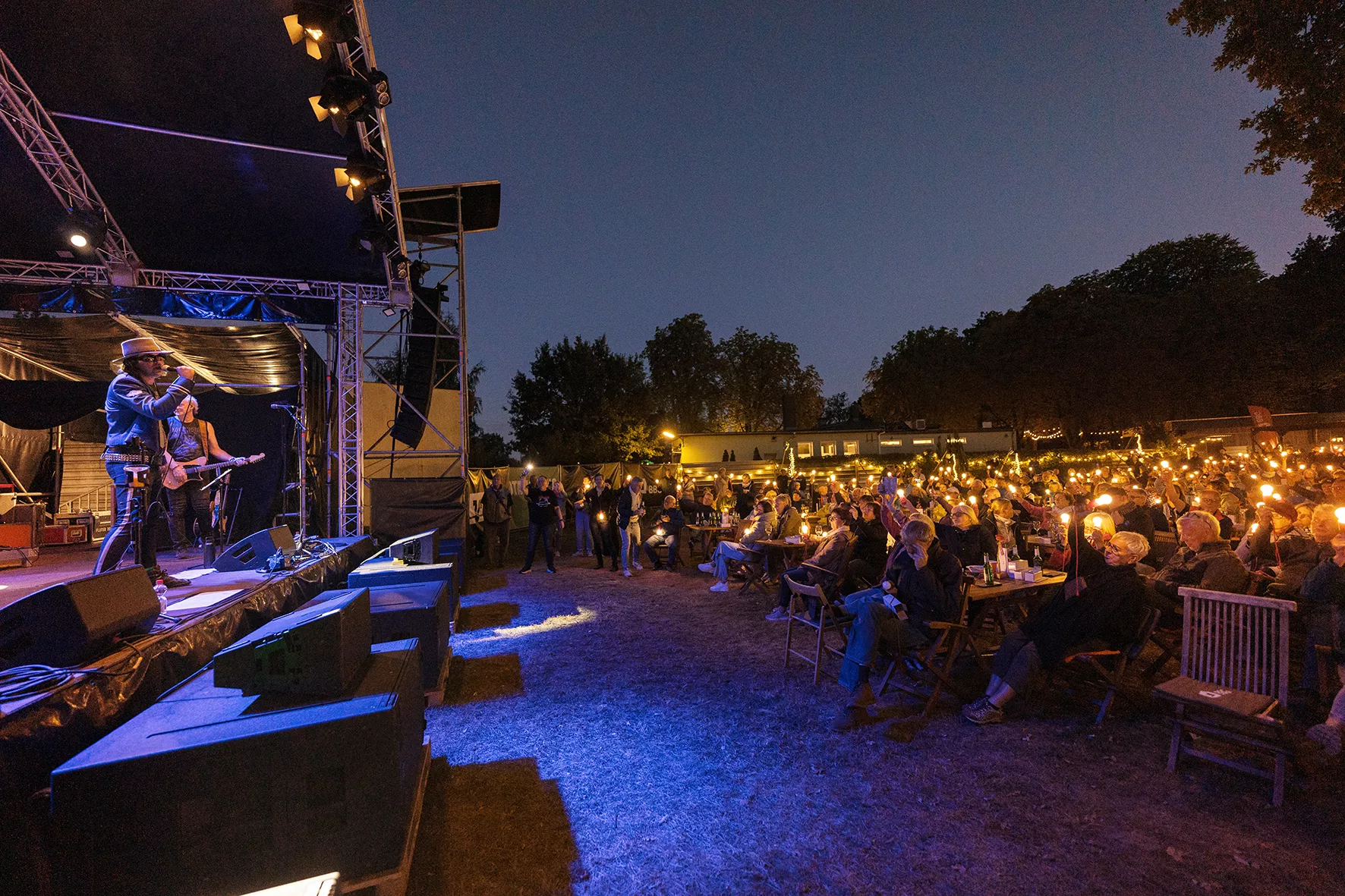 Eine Band spielt auf einer nächtlichen Freilichtbühne, während das Publikum leuchtende Lichter hält, die eine warme, festliche Atmosphäre schaffen.