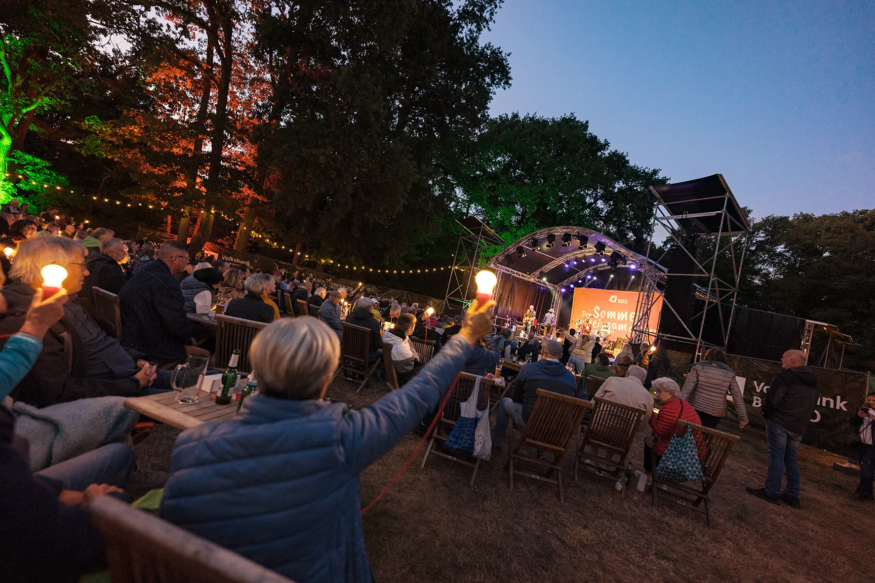 Die Besucher eines abendlichen Open-Air-Konzerts tragen Taschenlampen bei sich, während sie den Auftritt einer Band auf einer mit bunten Lichtern und Bäumen geschmückten Bühne verfolgen.