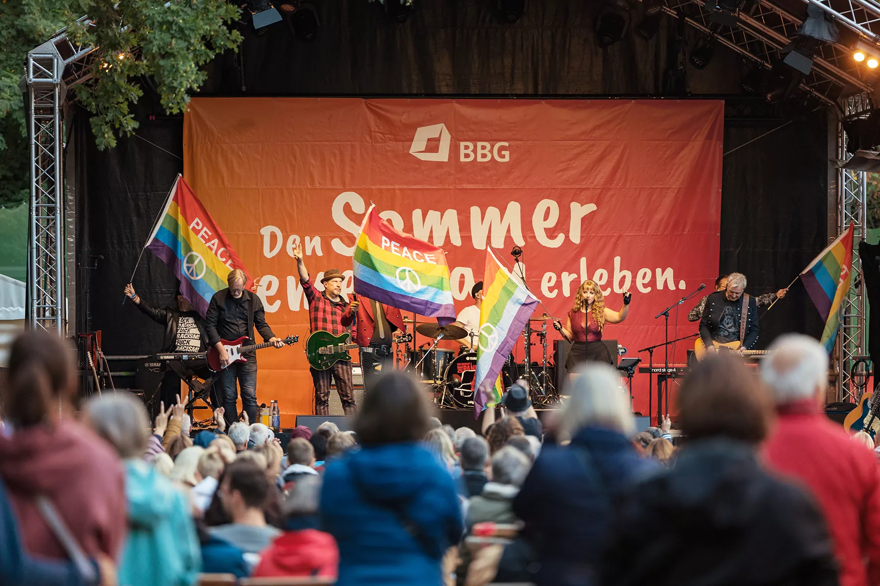 Eine Band spielt auf einer Open-Air-Bühne, während die Menschen regenbogenfarbene Friedensfahnen schwenken, um die Menge bei einer Sommerveranstaltung zu versammeln.