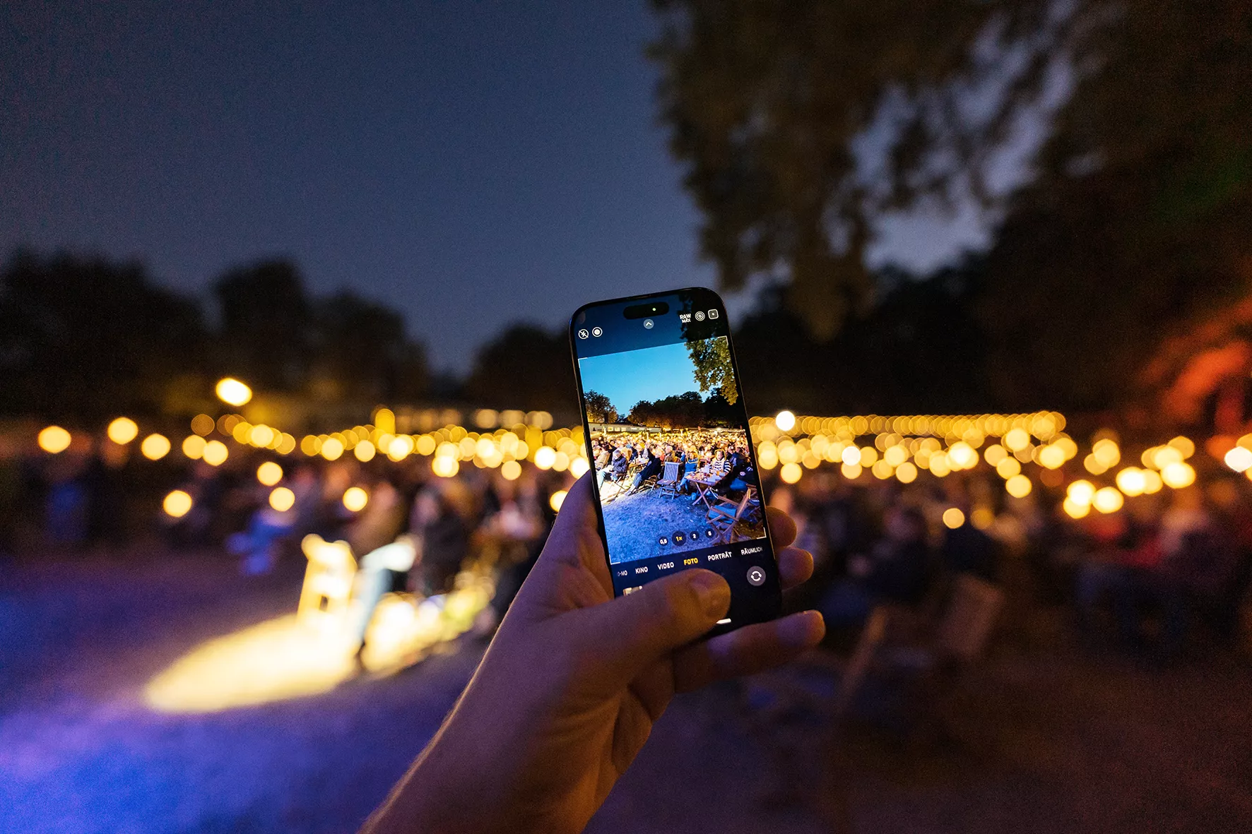 Eine Hand, die ein Telefon hält, nimmt eine Veranstaltung im Freien mit Lichterketten und sitzenden Menschen in der Abenddämmerung auf.