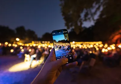 Eine Hand, die ein Telefon hält, nimmt eine Veranstaltung im Freien mit Lichterketten und sitzenden Menschen in der Abenddämmerung auf.