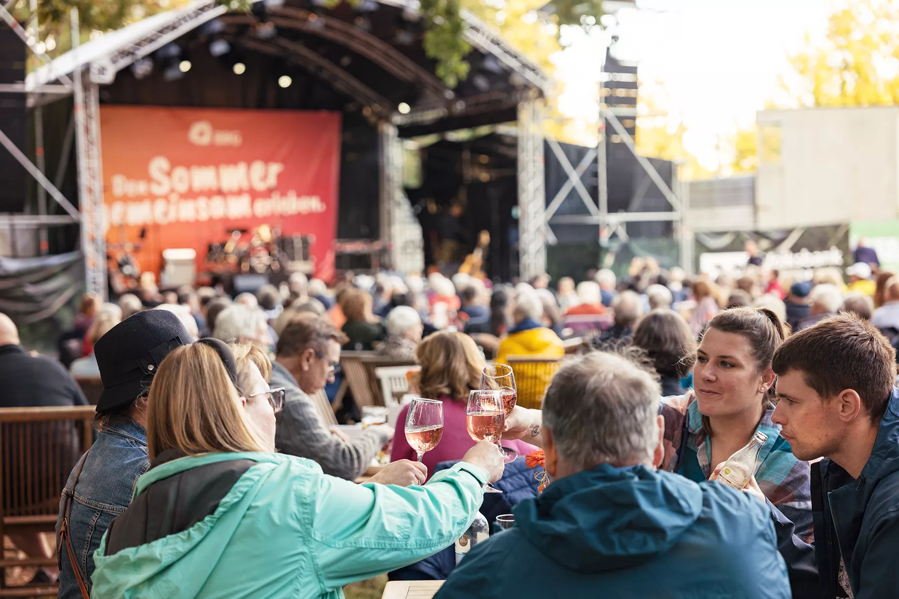 Menschen sitzen an Tischen im Freien und stoßen mit Gläsern auf ein energiegeladenes Konzert an, mit einer Bühne und Publikum im Hintergrund.