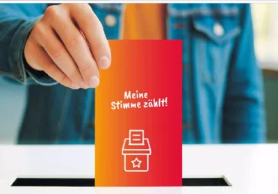 Eine Person wirft eine Karte mit der Aufschrift Meine Stimme zählt! in eine Wahlurne. Die Karte ist rot und orange mit einem Wahlsymbol.