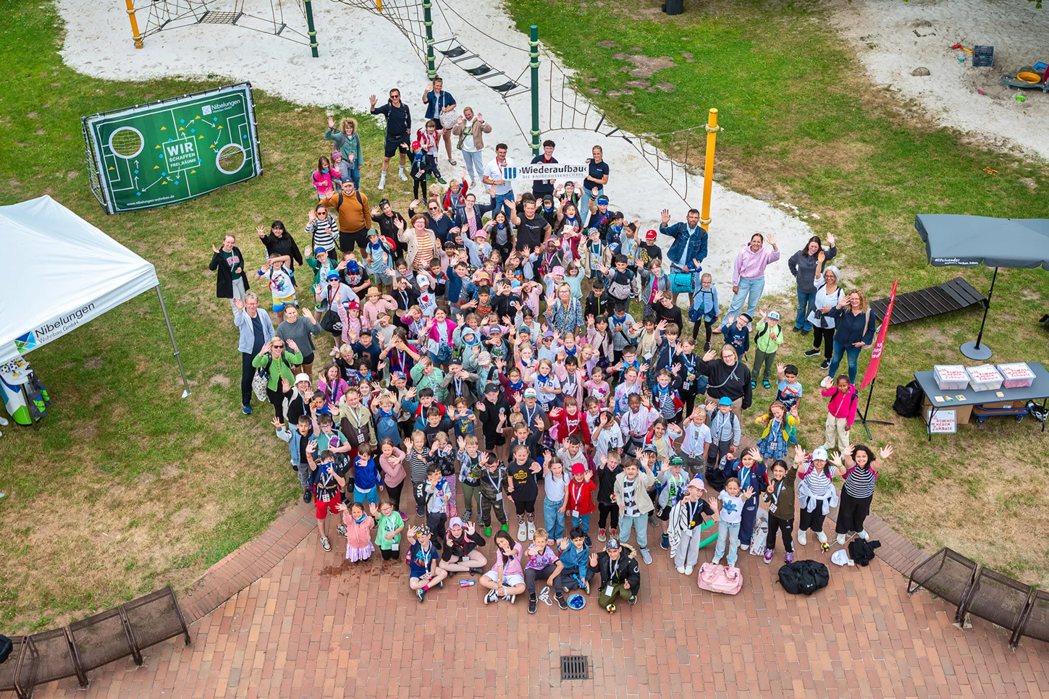 Eine große Gruppe von Kindern und Erwachsenen steht bei einer Veranstaltung im Freien auf einem Spielplatz zusammen, lächelt und winkt in die Kamera.