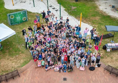Eine große Gruppe von Kindern und Erwachsenen steht bei einer Veranstaltung im Freien auf einem Spielplatz zusammen, lächelt und winkt in die Kamera.
