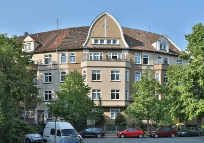 Ein beigefarbenes Wohnhaus mit rotem Ziegeldach, großen Fenstern, Bäumen davor und parkenden Autos entlang der Straße.