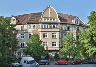 A beige-colored house with a red tiled roof, large windows, trees in front and parked cars along the street.